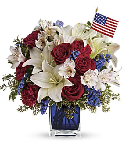 A patriotic pick to send a brave veteran, decorate your Fourth of July picnic, or celebrate Memorial Day. Lush red, white and blue flowers are presented in a deep blue glass cube vase along with an American flag.
Red roses, white asiatic lilies, white alstroemeria, blue statice and red carnations are gathered with green pitta negra, seeded eucalyptus and a small American flag in a cobalt blue glass cube.